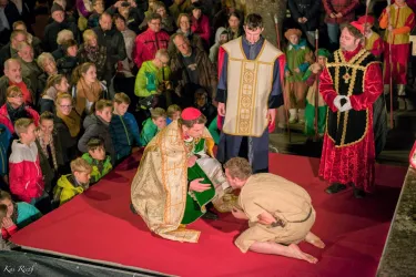 Eine lebhafte Aufführung mit Schauspielern in historischen Kostümen auf einer roten Bühne. Im Vordergrund kniet ein Darsteller vor einer weiteren Figur, während das Publikum zuschaut.