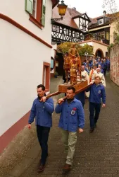 Eine Gruppe von Menschen trägt eine Statue durch eine enge Straße. Sie sind in traditionellen Kleidern gekleidet und wirken feierlich.