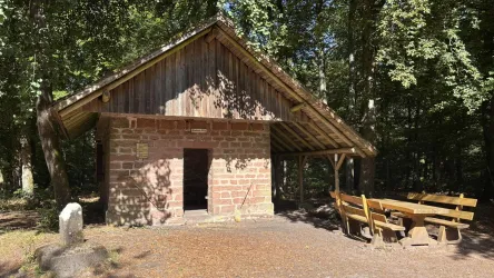 Eine rustikale Hütte im Wald mit einem überdachten Bereich. Draußen stehen mehrere Holzbänke und ein Tisch.