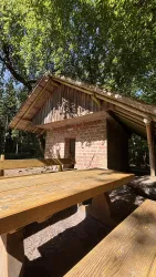 Eine gemütliche Hütte aus Stein und Holz unter einem Baumdach. Im Vordergrund steht ein holzfarbenen Tisch.