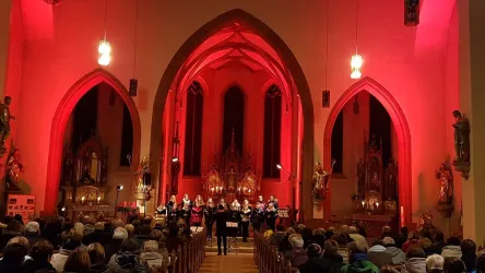 Eine Gruppe von Sängern tritt in einer festlich beleuchteten Kirche auf. Die Zuschauer sitzen in einem vollen Gottesdienstsaal unter roten Lichtern.
