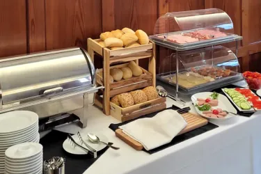 Ein Buffet mit verschiedenen Speisen, darunter Brötchen, Aufschnitt und frisches Gemüse. Es gibt auch Teller und Besteck für die Gäste.