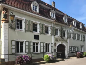 Ein traditionelles Gasthaus mit grünen Fensterläden und blühenden Blumen. Die Fassade ist hell und einladend, mit einem klaren blauen Himmel im Hintergrund.
