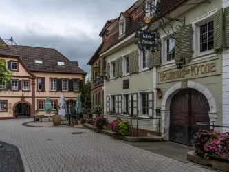 Eine malerische Straße mit alten Gebäuden und blühenden Pflanzen. Im Hintergrund ist eine Gaststätte mit dem Namen "Gaststätte Krone" zu sehen.
