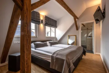Ein modernes Schlafzimmer mit Holzstrahlen und einem Doppelbett. Es gibt Fenster mit Vorhängen und Zugang zu einem Badezimmer.