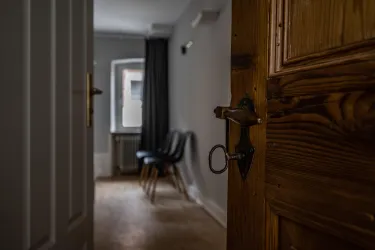 Eine geöffnete Holztür führt in einen hellen Raum mit einem Stuhl und einem Fenster. Die Wände sind neutral gestrichen, und der Boden ist aus Holz.