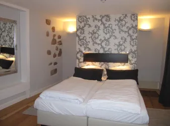 Ein helles Schlafzimmer mit einem Doppelbett und einem dekorativen Wandmuster. Ein Spiegel und sanfte Beleuchtung schaffen eine gemütliche Atmosphäre.