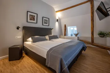 Ein modernes Schlafzimmer mit einem großen Doppelbett und Holzfußboden. An der Wand hängen schwarze Bilderrahmen, und es gibt ein Fenster, das natürlichem Licht hereinlässt.