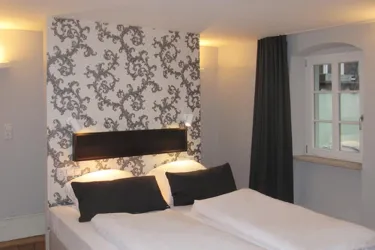 Ein modernes Schlafzimmer mit einem Doppelbett und eleganten Kissen. Die Wände sind mit einem schicken Muster dekoriert, und ein Fenster sorgt für Tageslicht.