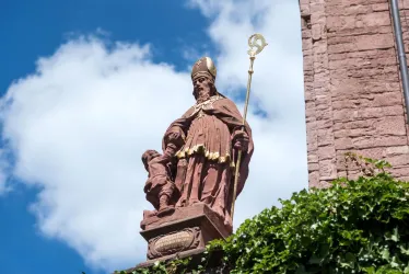 Eine Statue eines heiligen Bischofs mit einem Kind, die auf einem Gebäude steht. Im Hintergrund sind blaue Himmel und einige Wolken zu sehen.