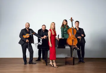 Eine Gruppe von fünf Musikern steht zusammen, wobei eine Frau in einem roten Kleid im Vordergrund hervorsticht. Im Hintergrund befinden sich ein Klavier und verschiedene Instrumente.