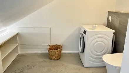 Ein heller und moderner Waschraum mit einer Waschmaschine, einem Wäschekorb und einem großen Fenster. Der Boden ist aus Beton und die Wände sind in neutralen Farben gehalten.