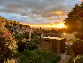 Ein malerischer Sonnenuntergang über einem Dorf, umgeben von bunten Gärten und Bäumen. Die Wolken reflektieren das warme Licht des Sonnenuntergangs.