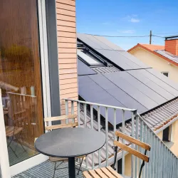 Ein moderner Balkon mit einem runden Tisch und zwei Stühlen. Im Hintergrund sind Solarpanels auf einem Dach zu sehen.