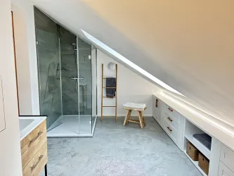 Ein modernes Badezimmer in einem Dachgeschoss mit einer gläsernen Dusche und minimalistischen Möbeln. Die Wände sind hell und der Raum wirkt geräumig und einladend.