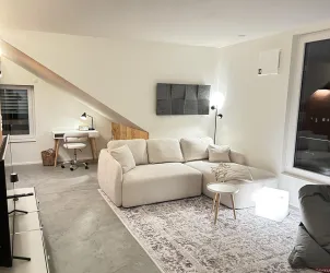 Ein modernes Wohnzimmer mit einem gemütlichen, hellen Sofa und einem kleinen Tisch. An der Wand befindet sich ein Schreibtisch mit einem Stuhl und einem Fenster, das viel Licht hereinlässt.