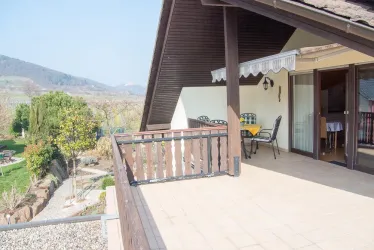 Ein Balkon mit Blick auf eine grüne Landschaft und Berge im Hintergrund. Tische und Stühle laden zum Entspannen ein.