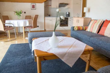 Ein gemütliches Wohnzimmer mit einem blauen Teppich und einem Sofa. Auf dem Tisch steht eine Vase mit Blumen und im Hintergrund ist ein Esstisch zu sehen.