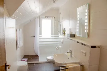 Ein modernes Badezimmer mit hellem Fliesenboden und einer Duschkabine. Es gibt ein Fenster, Handtücher und eine elegante Waschbeckenanordnung.