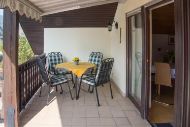 Ein gemütlicher Balkon mit einem Tisch und vier Stühlen. Auf dem Tisch steht eine kleine Blume, und die Aussicht scheint einladend zu sein.