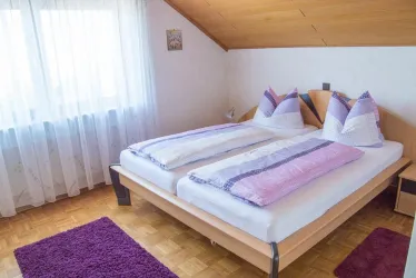 Ein gemütliches Schlafzimmer mit einem Doppelbett und bunten Kissen. Das Zimmer hat große Fenster, die viel Tageslicht hereinlassen.