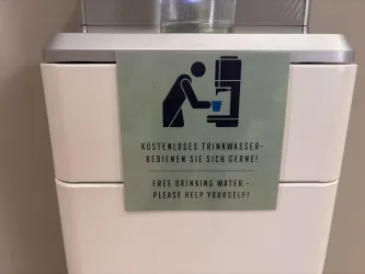 Ein Wasserspender mit einem Schild, das zum kostenlosen Trinken einlädt. Der Hinweis enthält Anweisungen auf Deutsch und Englisch.