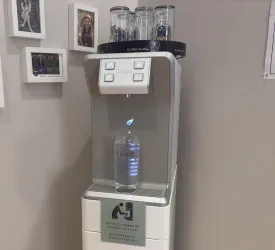 Ein moderner Wasserspender in einem Raum mit Fotos an der Wand. Der Spender hat eine klare Flasche und zeigt eine leuchtende Anzeige.
