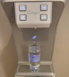 Ein moderner Wasserspender mit leuchtenden Tasten und einem Glas darunter. Das Glas ist teilweise mit klarem Wasser gefüllt.