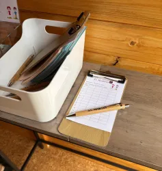 Ein weißer Behälter mit Dokumenten steht auf einem Tisch. Daneben liegt eine beschreibbare Klemmbrett mit einem Stift.