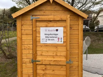 Eine kleine Holzschutzhütte mit einem Schild darauf. Das Schild weist darauf hin, dass Minigolf-Schläger verfügbar sind.