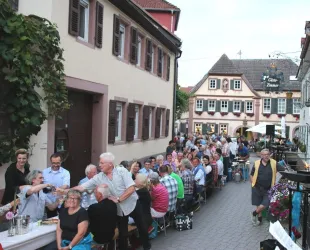 Eine lange Tafel mit vielen Menschen, die gemeinsam essen und trinken, steht auf einer Straße. Die Umgebung zeigt charmante, historische Gebäude und eine gemütliche Atmosphäre.