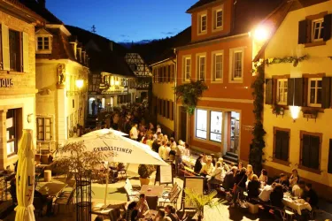 Eine charmante Gasse mit beleuchteten Häusern und vielen Menschen, die an Tischen sitzen. Die Atmosphäre ist abends lebhaft und einladend.