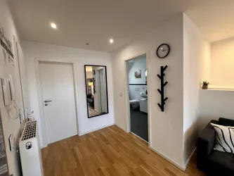 Ein heller Flur mit Holzfußboden und weißen Wänden. An der Wand hängt ein Spiegel und ein Wandregal.