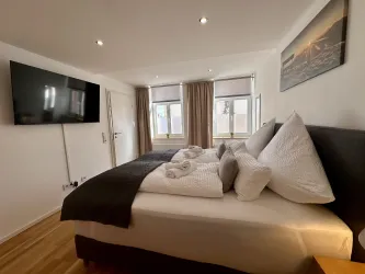 Ein modernes Schlafzimmer mit einem großen Bett und mehreren Kissen. Es gibt einen Flachbildfernseher und Fenster mit Vorhängen.