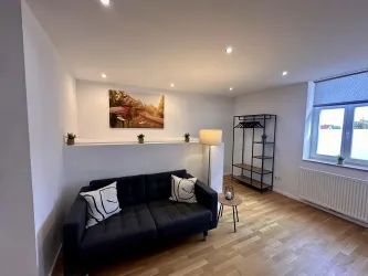 Ein modernes Wohnzimmer mit einem schwarzem Sofa und einer stylischen Lampe. An der Wand hängt ein Bild, während ein offenes Regal im Raum steht.