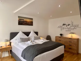 Ein modernes Schlafzimmer mit einem Doppelbett, stilvollen Kissen und einer warmen Holzkommode. An der Wand hängt ein Bild einer Stadtlandschaft sowie inspirierende Zitate über die Natur.