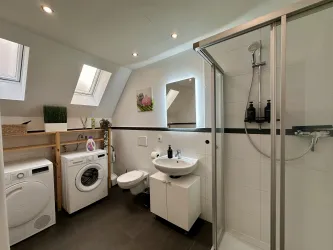 Ein modernes Badezimmer mit einer Duschkabine, Waschmaschine und Waschbecken. Das Zimmer hat Tageslicht durch zwei Dachfenster und dezente Dekoration.
