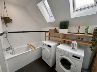 Ein modernes Badezimmer mit einer Badewanne und zwei Waschmaschinen. Die Wände sind hell, und es gibt dekorative Pflanzen sowie praktische Regale.