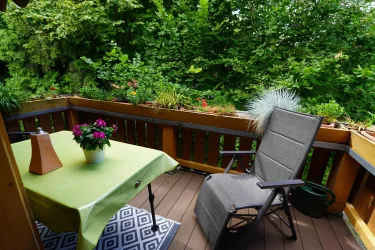 Ein gemütlicher Balkon mit einem Tisch und einem Stuhl. Auf dem Tisch steht eine Pflanze und der Balkon ist von üppigem Grün umgeben.