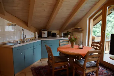 Eine gemütliche Küche mit Holzbalkendecke und blauen Küchenschränken. Ein runder Tisch mit Stühlen und einer Blumenvasen auf der Tischplatte.