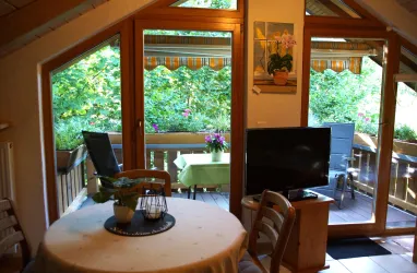 Ein gemütlicher Raum mit einem runden Tisch und einem Fernseher. Durch große Fenster sieht man einen blühenden Balkon mit einem grünen Ausblick.