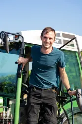 Ein lächelnder Mann steht an einem Traktor und schaut in die Kamera. Es ist ein sonniger Tag mit klarem Himmel.