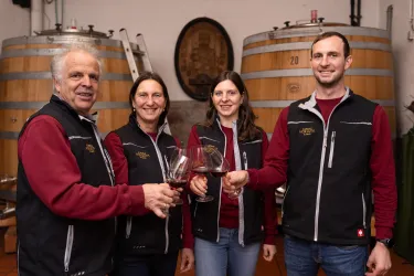 Eine Gruppe von vier Personen in einem Weinkeller hält Gläser mit Rotwein. Im Hintergrund sind große Holzfässer zu sehen.