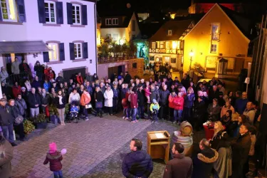 Eine große Menschenmenge versammelt sich auf einem Platz bei Nacht. Umgeben von festlich beleuchteten Häusern, scheinen die Menschen eine Veranstaltung zu genießen.