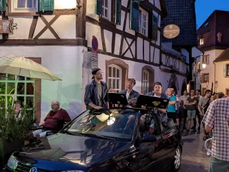 Eine Gruppe von Musikern steht auf einem Auto und spielt Musik in einer belebten Straße. Im Hintergrund sind Zuschauer und historische Häuser zu sehen.