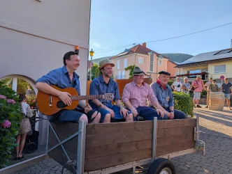 Vier Männer sitzen in einem Wagen und spielen Musik. Im Hintergrund sind einige Häuser und Menschen zu sehen.