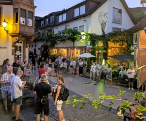 Eine lebhafte Straße mit vielen Menschen in einem historischen Stadtviertel. Die Umgebung ist von Pflanzen und warmem Licht beleuchtet.