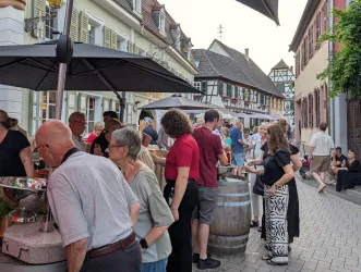 Eine belebte Straße mit Menschen, die an einem Marktstand stehen. Es gibt Stände und traditionelle Architektur im Hintergrund.