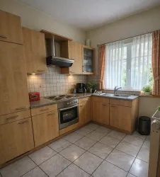 Eine moderne Küche mit Holzschränken und einer hellen Arbeitsfläche. Sie verfügt über einen Herd, eine Spüle und viel Tageslicht, das durch die Fenster strömt.