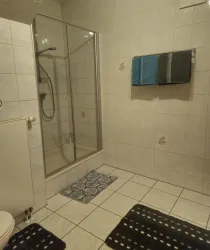 Ein modernes Badezimmer mit einer gläsernen Dusche und weißen Fliesen. Handtücher hängen an der Wand und es liegen Teppiche auf dem Boden.